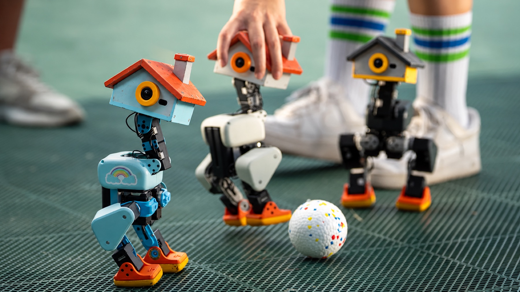 Three NANOPONS robots on a green court with a ball, a person standing behind them